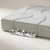 Lănțișor argint 925 rodiat personalizat cu numele Antonia decupat în scriere cursivă elegantă cu coadă caligrafică pe placă de beton gri, lanț subțire alb-platinat