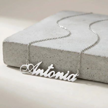 Lănțișor argint 925 rodiat personalizat cu numele Antonia decupat în scriere cursivă elegantă cu coadă caligrafică pe placă de beton gri, lanț subțire alb-platinat