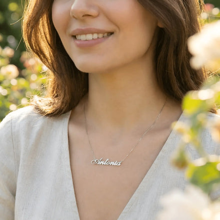Damă tânără purtând lănțișor argint 925 rodiat cu numele Antonia script, peste top crem de in cu decolteu V, lumină naturală exterior cu trandafiri