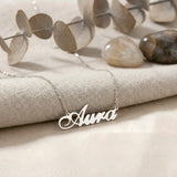 Lănțișor argint 925 rodiat personalizat cu numele Aura decupat în scriere cursivă elegantă cu A larg și coadă caligrafică pe textil bej cu eucalipt și pietre, lanț subțire alb-platinat