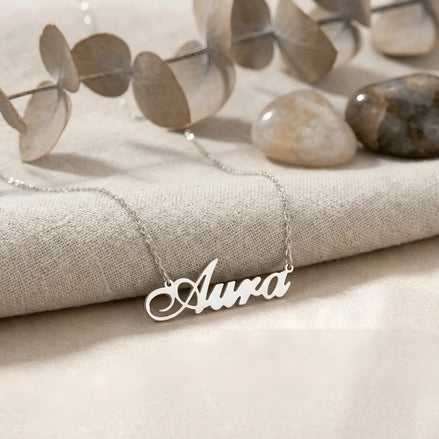 Lănțișor argint 925 rodiat personalizat cu numele Aura decupat în scriere cursivă elegantă cu A larg și coadă caligrafică pe textil bej cu eucalipt și pietre, lanț subțire alb-platinat