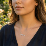 Damă tânără purtând lănțișor argint 925 rodiat cu numele Aura script, peste top bleumarin cu decolteu V, lumină naturală exterior cu trandafiri