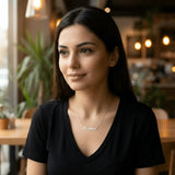 Damă tânără purtând lănțișor argint 925 rodiat cu numele Beatrice script, peste t-shirt negru cu decolteu V, cafenea cu lumină caldă și plante