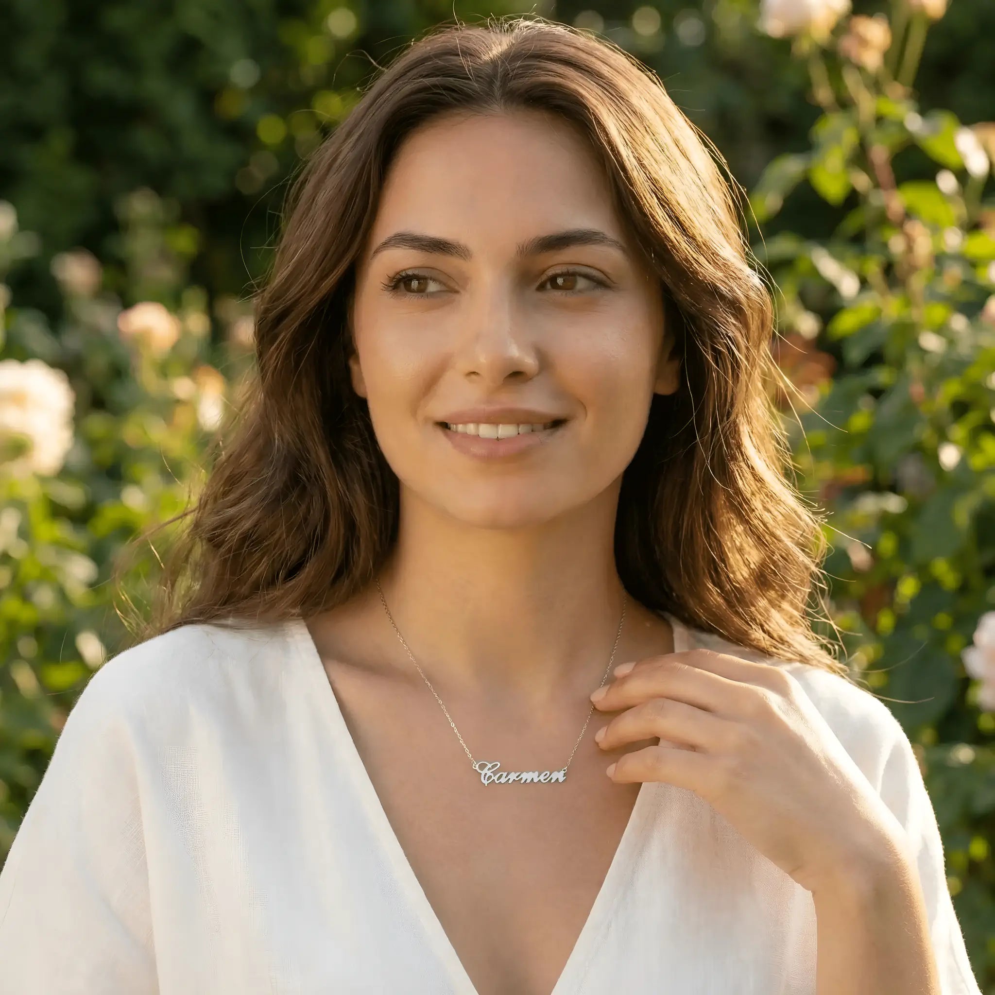 Damă tânără purtând lănțișor argint 925 rodiat cu numele Carmen script, peste top alb cu decolteu V, lumină naturală exterior cu trandafiri