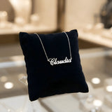 Lănțișor argint 925 rodiat cu numele Claudia, pandantiv decupat 42x10 mm font cursiv inițiala C deschisă, lanț anchor 46 cm pe pernuță velvet bleumarin într-o vitrină de bijuterii