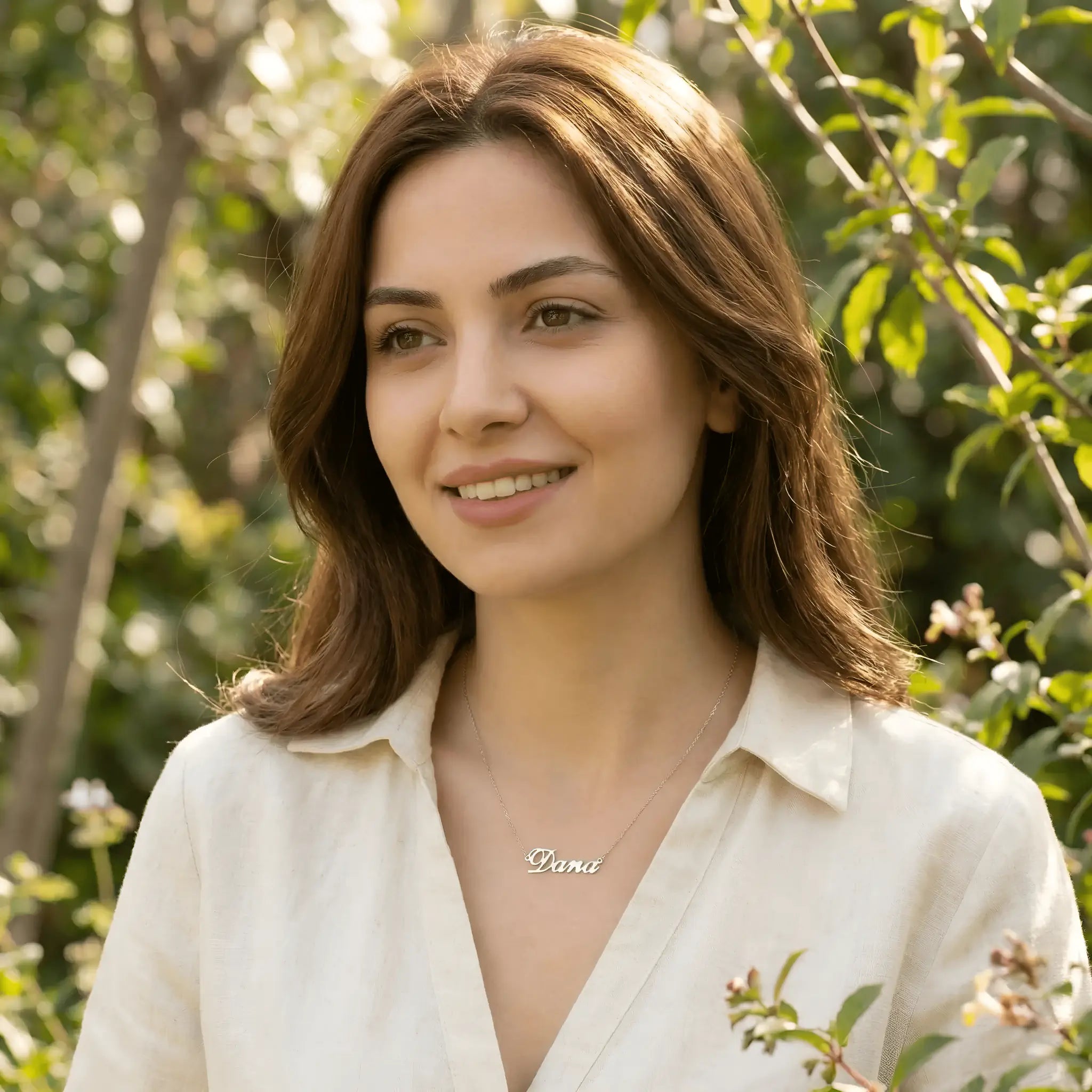 Damă tânără purtând lănțișor argint 925 rodiat cu numele Dana script, peste cămașă crem de in cu primii nasturi deschiși, lumină caldă exterior cu vegetație verde