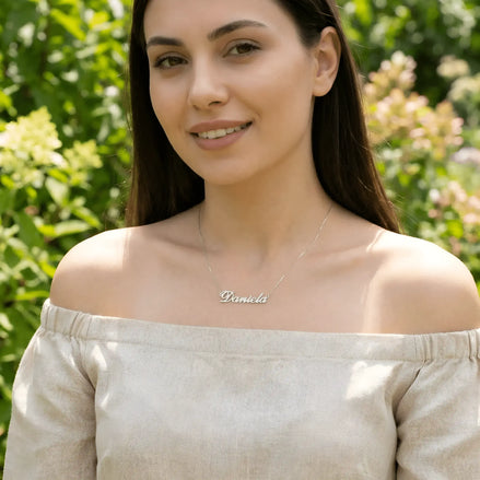 Damă tânără purtând lănțișor argint 925 rodiat cu numele Daniela script, peste top off-shoulder de in bej, lumină naturală exterior cu vegetație