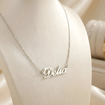 Lănțișor argint 925 rodiat personalizat cu numele Delia decupat în scriere cursivă elegantă cu D buclat pe drapaj de mătase crem cu bust display, lanț subțire alb-platinat