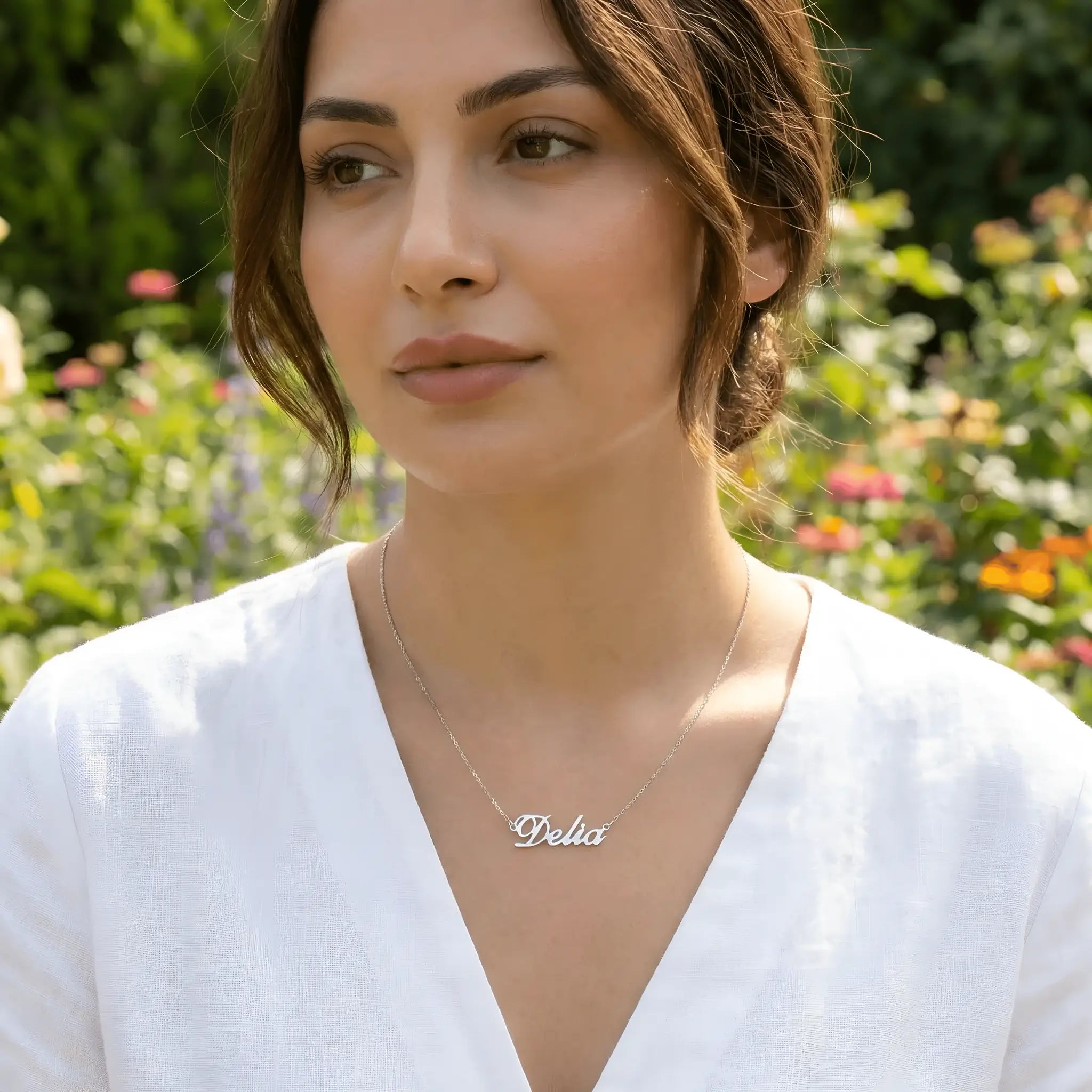 Damă tânără purtând lănțișor argint 925 rodiat cu numele Delia script, peste bluză albă de in cu decolteu V, lumină naturală exterior cu flori colorate