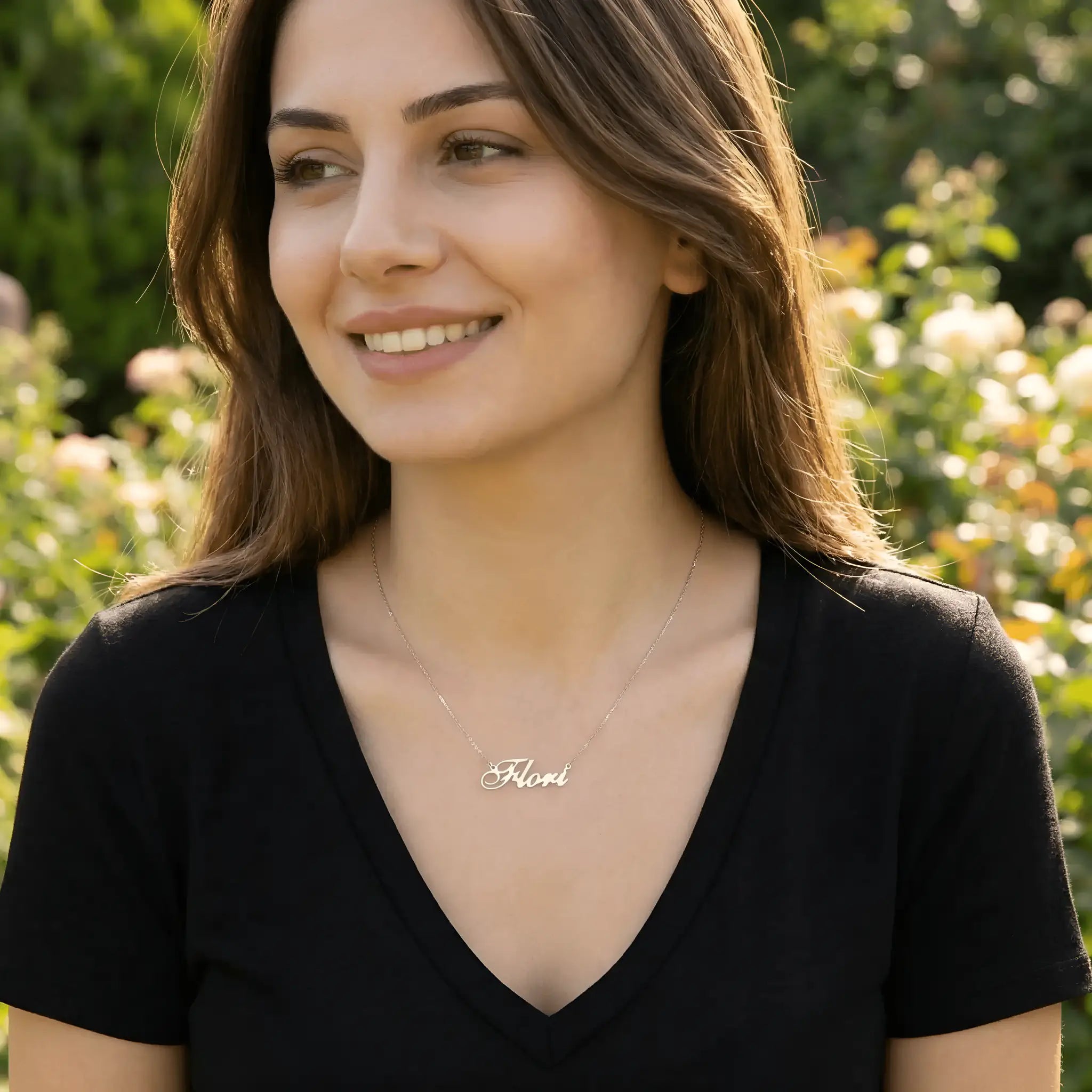 Damă tânără purtând lănțișor argint 925 rodiat cu numele Flori script, peste t-shirt negru cu decolteu V adânc, lumină naturală exterior cu trandafiri