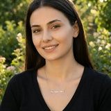 Damă tânără purtând lănțișor argint 925 rodiat cu numele Georgiana script, peste top negru cu decolteu V, lumină naturală exterior cu vegetație