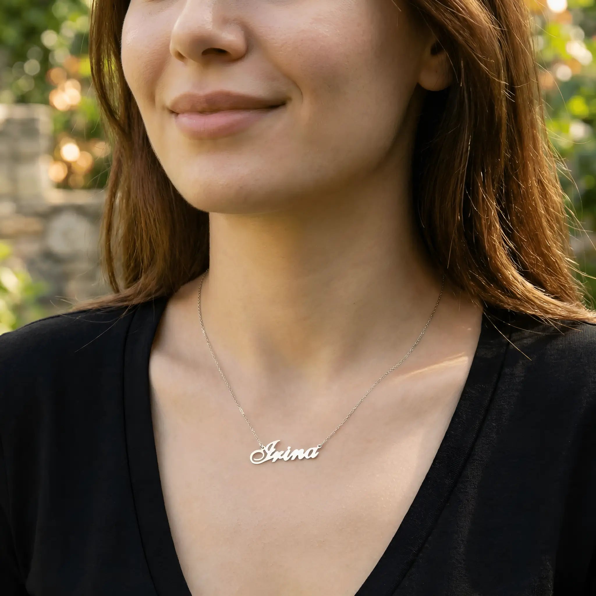 Damă tânără purtând lănțișor argint 925 rodiat cu numele Irina script, peste top negru cu decolteu V, lumină naturală exterior cu vegetație și zid de piatră