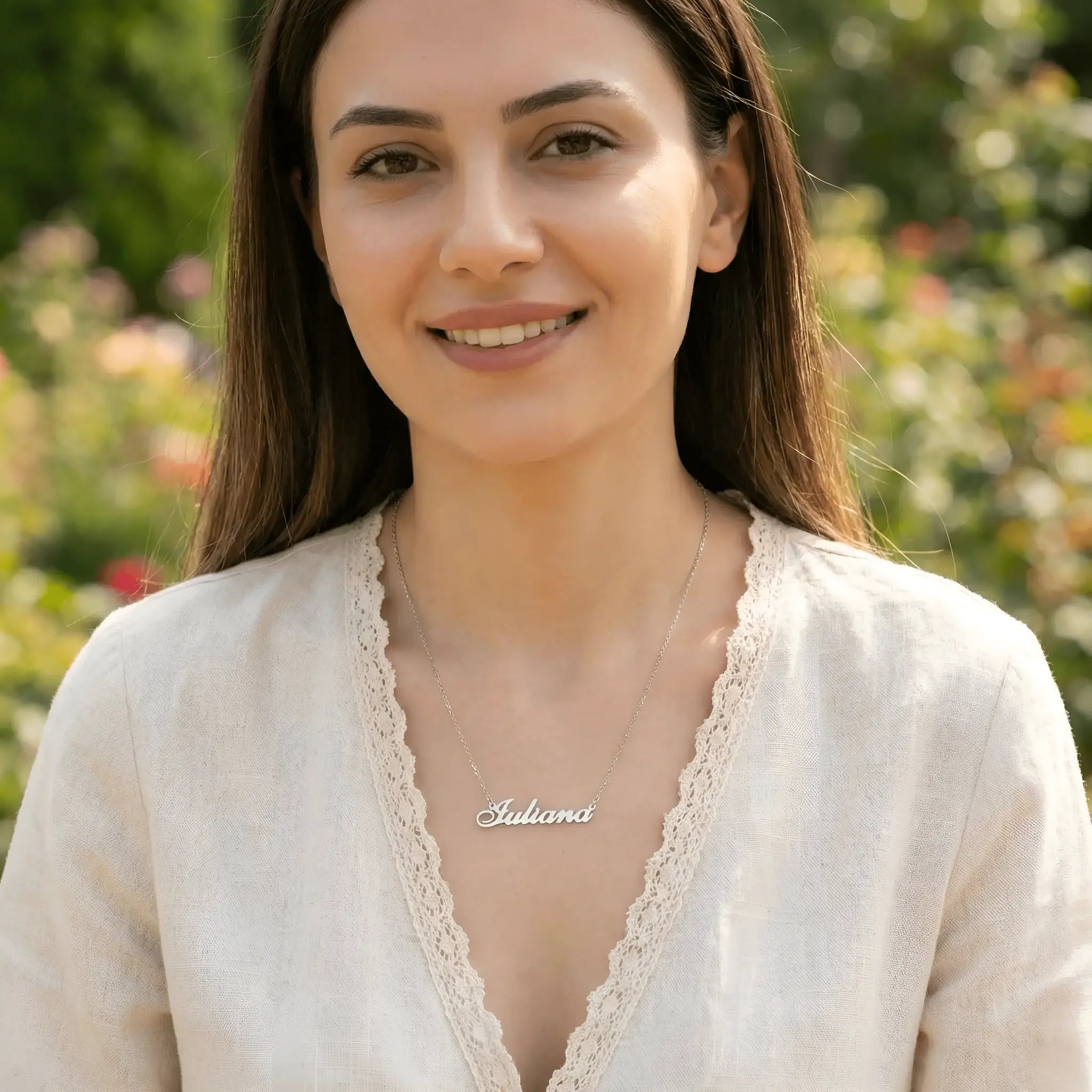 Damă tânără purtând lănțișor argint 925 rodiat cu numele Iuliana script, peste bluză albă cu dantelă cu decolteu V, lumină naturală exterior cu vegetație și flori