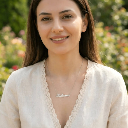 Damă tânără purtând lănțișor argint 925 rodiat cu numele Iuliana script, peste bluză albă cu dantelă cu decolteu V, lumină naturală exterior cu vegetație și flori