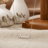 Lănțișor argint 925 rodiat personalizat cu numele Larisa decupat în scriere cursivă elegantă cu L larg pe textil bej cu vase ceramice albe și eucalipt, lanț subțire alb-platinat