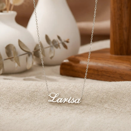 Lănțișor argint 925 rodiat personalizat cu numele Larisa decupat în scriere cursivă elegantă cu L larg pe textil bej cu vase ceramice albe și eucalipt, lanț subțire alb-platinat