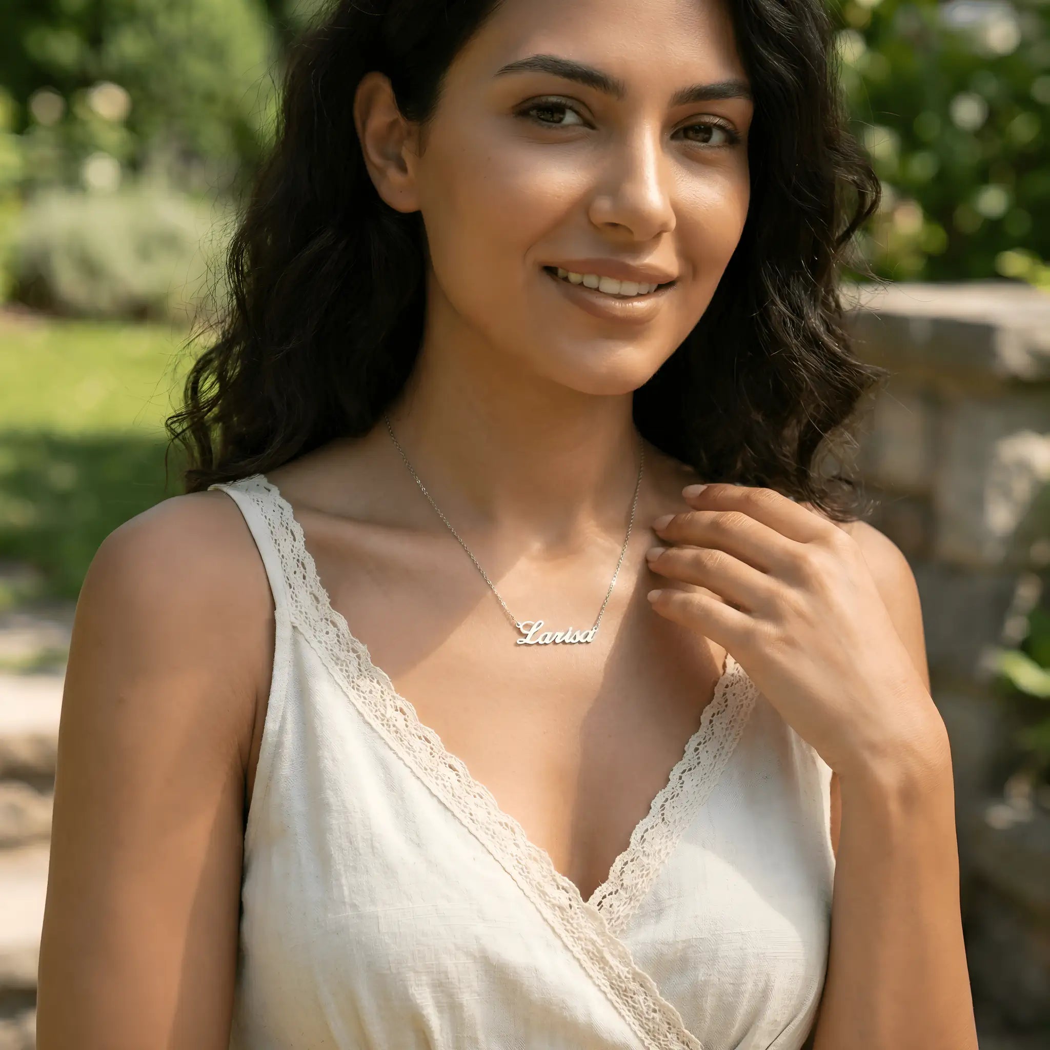 Damă tânără purtând lănțișor argint 925 rodiat cu numele Larisa script, peste top alb cu V wrap și dantelă, lumină naturală exterior cu vegetație și zid de piatră
