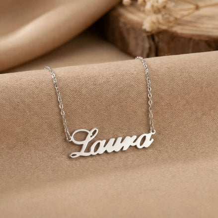 Lănțișor argint 925 rodiat personalizat cu numele Laura decupat în scriere cursivă elegantă cu L larg pe textil bej cu suport lemn rotund, lanț subțire alb-platinat