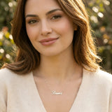 Damă tânără purtând lănțișor argint 925 rodiat cu numele Laura script, peste pulover crem fin de cașmir cu decolteu V, lumină naturală exterior toamnă