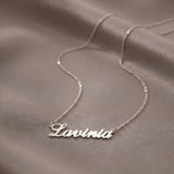 Lănțișor argint 925 rodiat personalizat cu numele Lavinia decupat în scriere cursivă elegantă cu L larg pe drapaj satin maroniu mătăsos, lanț subțire alb-platinat