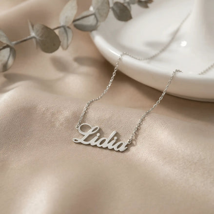 Lănțișor argint 925 rodiat personalizat cu numele Lidia decupat în scriere cursivă elegantă cu L larg pe drapaj satin roz cu eucalipt, lanț subțire alb-platinat