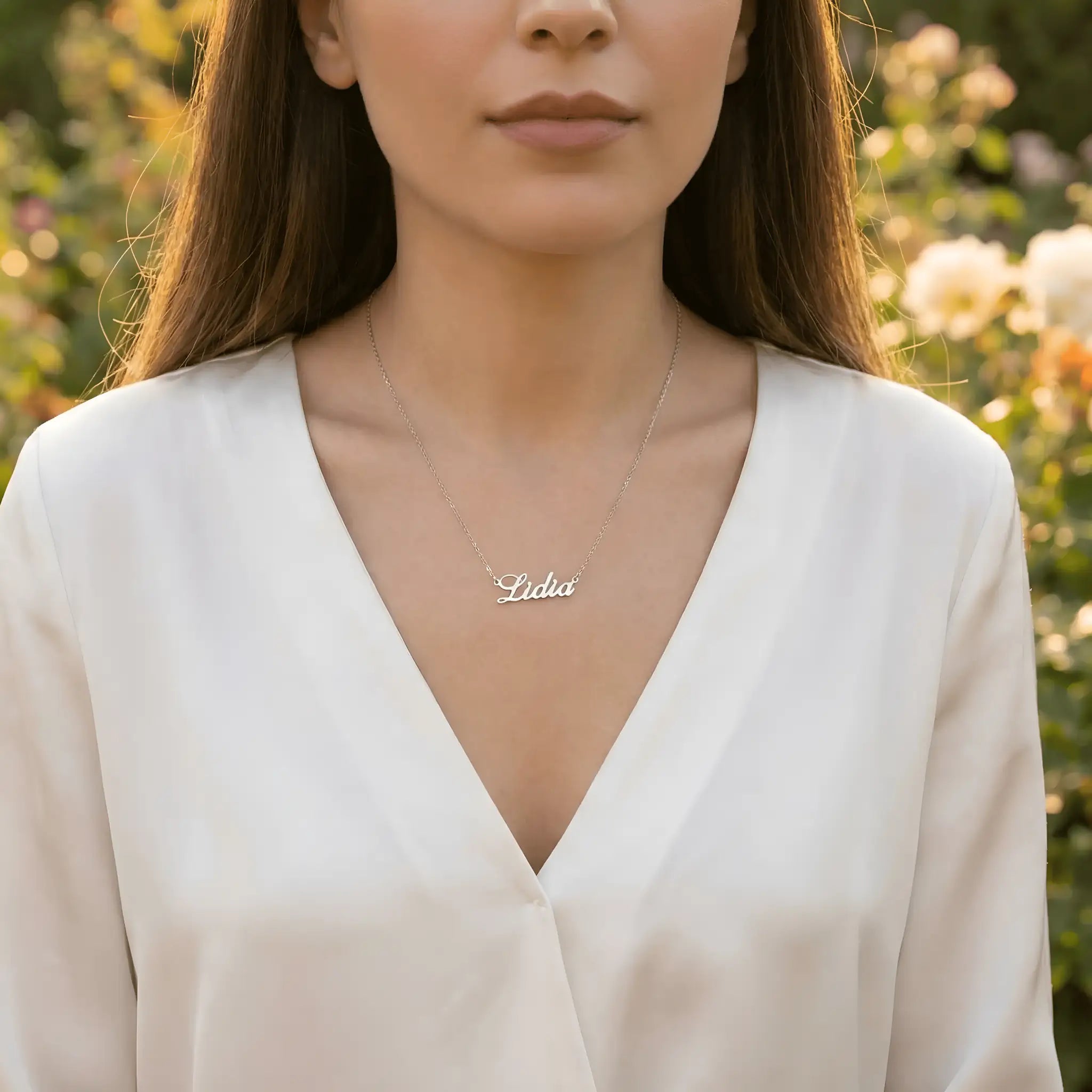 Damă tânără purtând lănțișor argint 925 rodiat cu numele Lidia script, peste bluză albă cu V wrap, lumină naturală exterior cu trandafiri pastel
