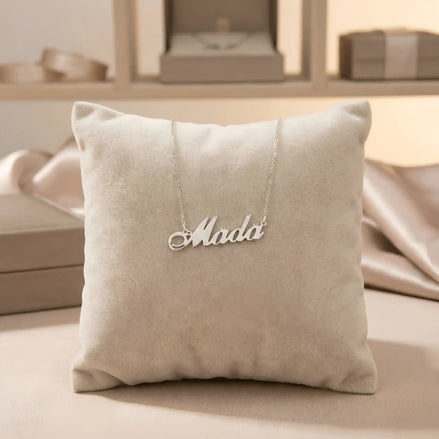 Lănțișor argint 925 rodiat personalizat cu numele Mada decupat în scriere cursivă elegantă cu M cu coadă caligrafică pe pernuță bej display velvet, lanț subțire alb-platinat
