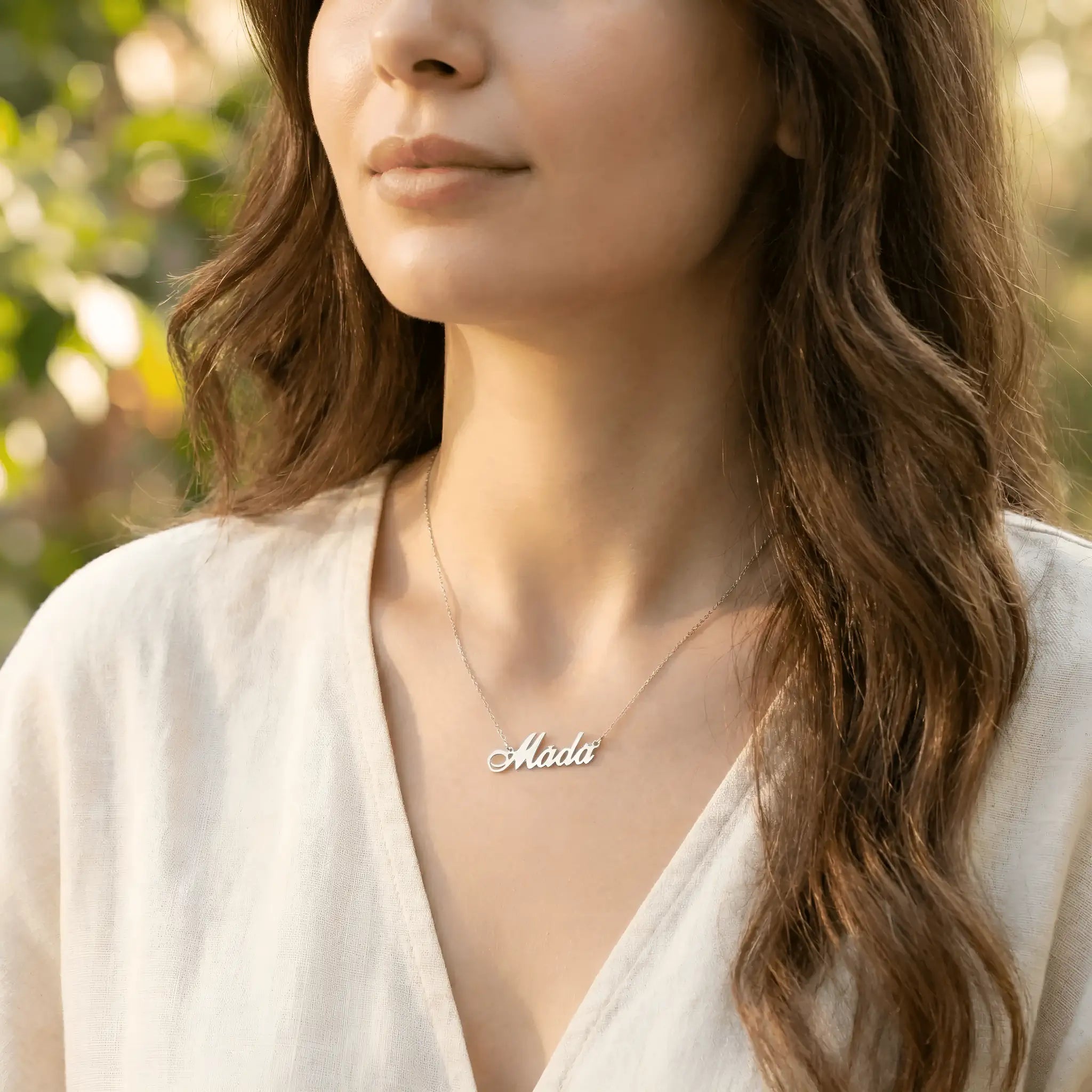 Damă tânără purtând lănțișor argint 925 rodiat cu numele Mada script, peste bluză albă cu decolteu V wrap, lumină naturală exterior cu vegetație