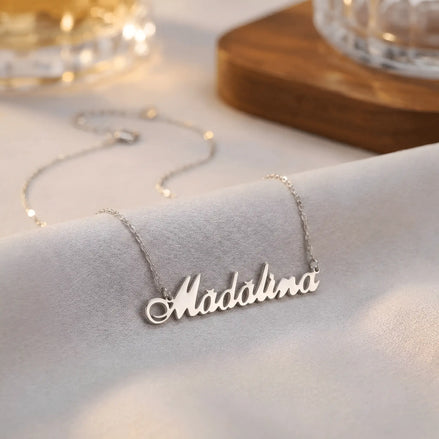 Lănțișor argint 925 rodiat cu numele Madalina, pandantiv decupat font cursiv italic legat, lanț anchor cu extender pe textil ivory mat, lângă obiecte de cristal