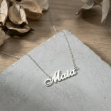 Lănțișor argint 925 rodiat personalizat cu numele Maia decupat în scriere cursivă elegantă cu M cu coadă caligrafică pe stâncă gri pe lemn cu frunze uscate, lanț subțire alb-platinat