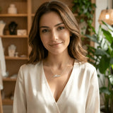 Damă tânără purtând lănțișor argint 925 rodiat cu numele Maia script, peste bluză albă de mătase cu V wrap, lumină caldă interior cu plante și mobilier de lemn