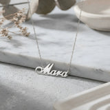 Lănțișor argint 925 rodiat cu numele Mara, pandantiv decupat 31x10 mm font cursiv inițiala M cu curbă dublă, lanț anchor 46 cm pe blat de marmură albă cu vase ceramice și ramură uscată
