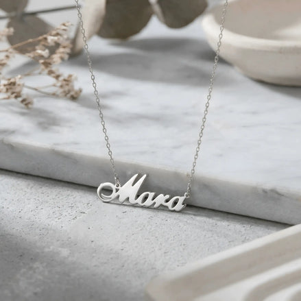 Lănțișor argint 925 rodiat cu numele Mara, pandantiv decupat 31x10 mm font cursiv inițiala M cu curbă dublă, lanț anchor 46 cm pe blat de marmură albă cu vase ceramice și ramură uscată