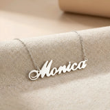 Lănțișor argint 925 rodiat personalizat cu numele Monica decupat în scriere cursivă elegantă cu M larg pe textil bej, lanț subțire alb-platinat