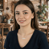 Damă tânără purtând lănțișor argint 925 rodiat cu numele Oana script, peste t-shirt bleumarin cu decolteu V, lumină caldă cafenea rustică cu mobilier de lemn