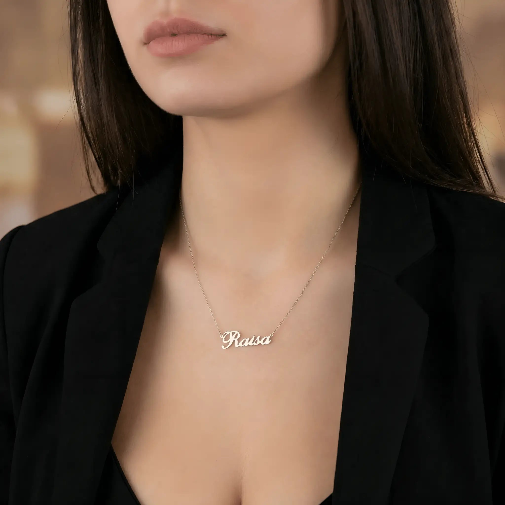 Damă cu păr lung negru purtând lănțișor argint 925 rodiat Raisa pe stern între reverele unui sacou negru, interior elegant ambient cald