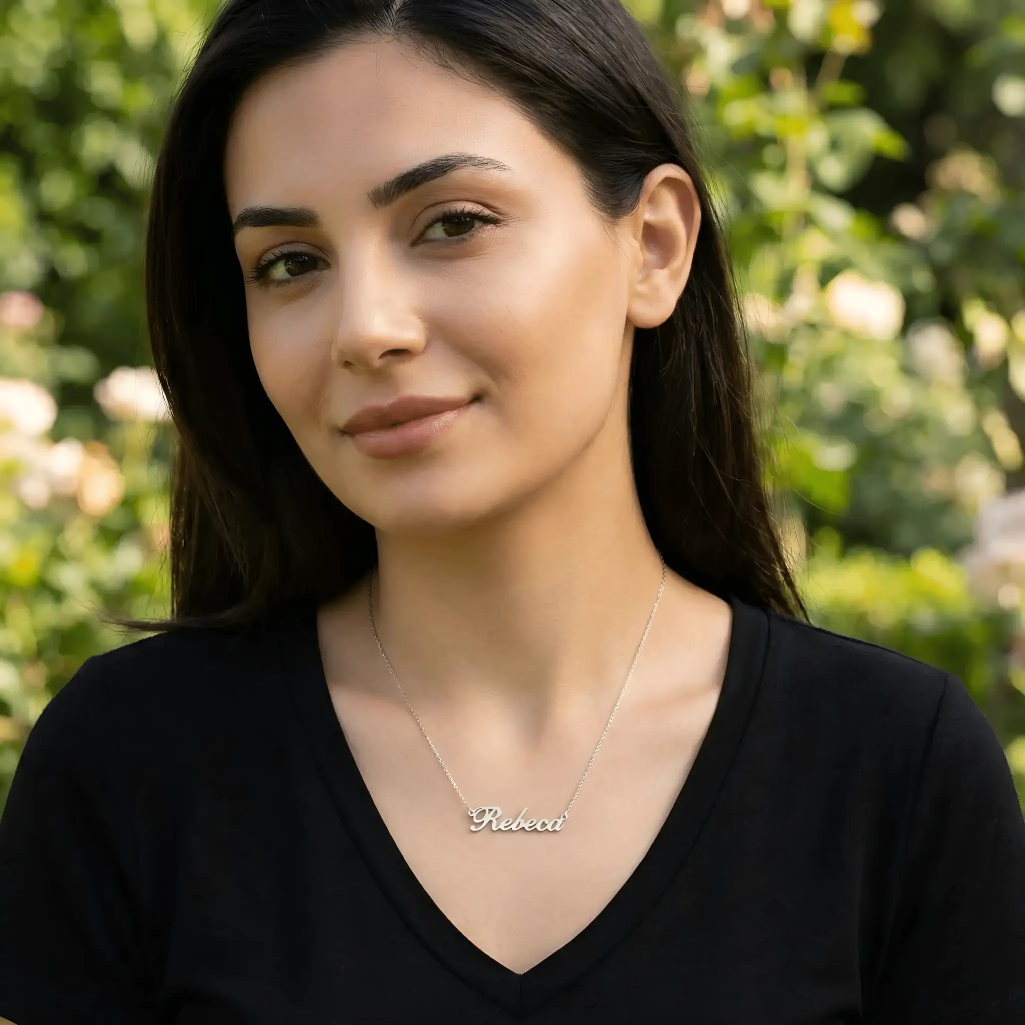 Damă tânără purtând lănțișor argint 925 rodiat cu numele Rebeca script, peste top negru cu decolteu V, lumină naturală exterior cu trandafiri