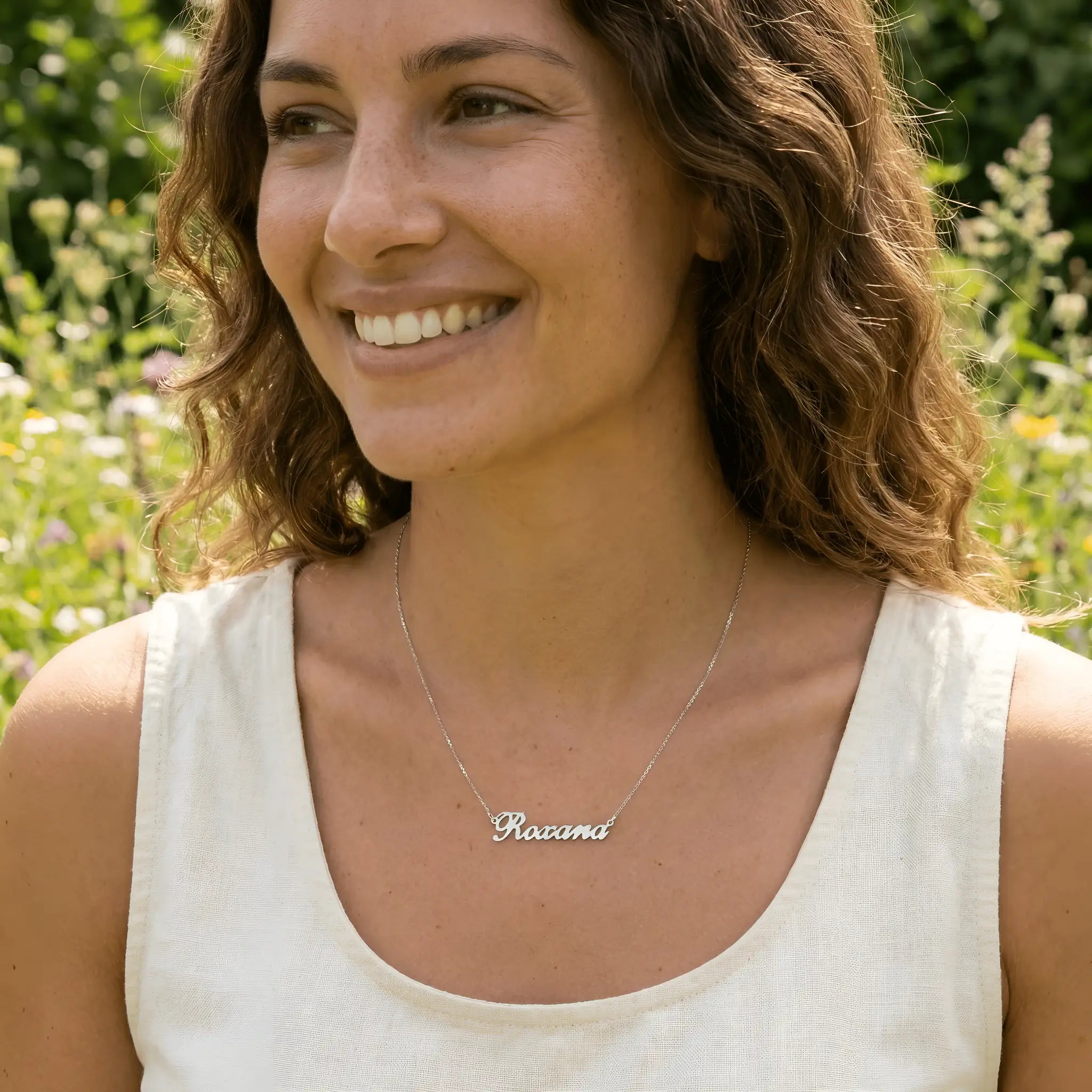 Damă tânără purtând lănțișor argint 925 rodiat cu numele Roxana script, peste top alb fără mâneci, lumină naturală exterior cu vegetație