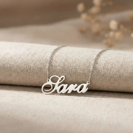 Lănțișor argint 925 rodiat personalizat cu numele Sara decupat în scriere cursivă elegantă cu S ondulat pe textil bej cu ramuri uscate, lanț subțire alb-platinat