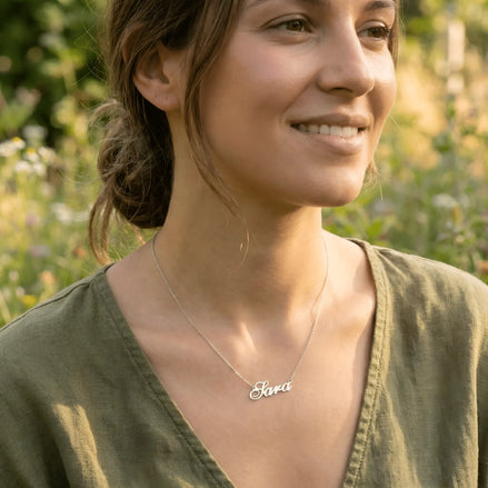 Damă tânără purtând lănțișor argint 925 rodiat cu numele Sara script, peste top de in kaki cu decolteu V, lumină naturală exterior cu wildflowers