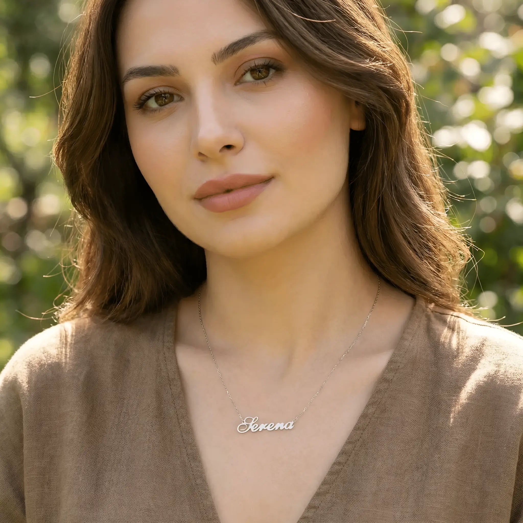 Damă tânără purtând lănțișor argint 925 rodiat cu numele Serena script, peste top maro brun de in cu decolteu V, lumină naturală exterior cu vegetație
