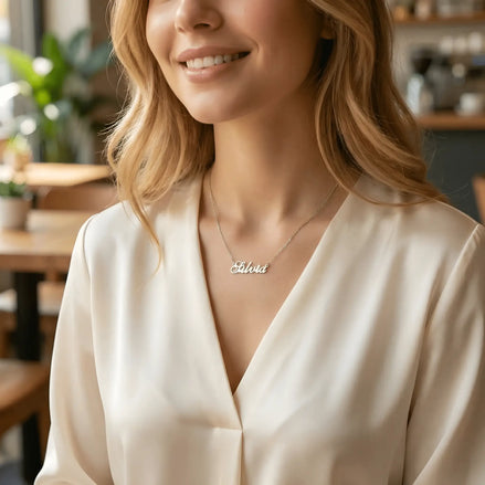 Damă tânără purtând lănțișor argint 925 rodiat cu numele Silvia script, peste bluză albă de mătase cu decolteu V, lumină caldă cafenea cu plante