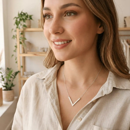 Damă tânără purtând lănțișor argint 925 rodiat cu pandantiv V geometric peste cămașă bej cu decolteu V, dublu V efect editorial, interior cozy cu plante